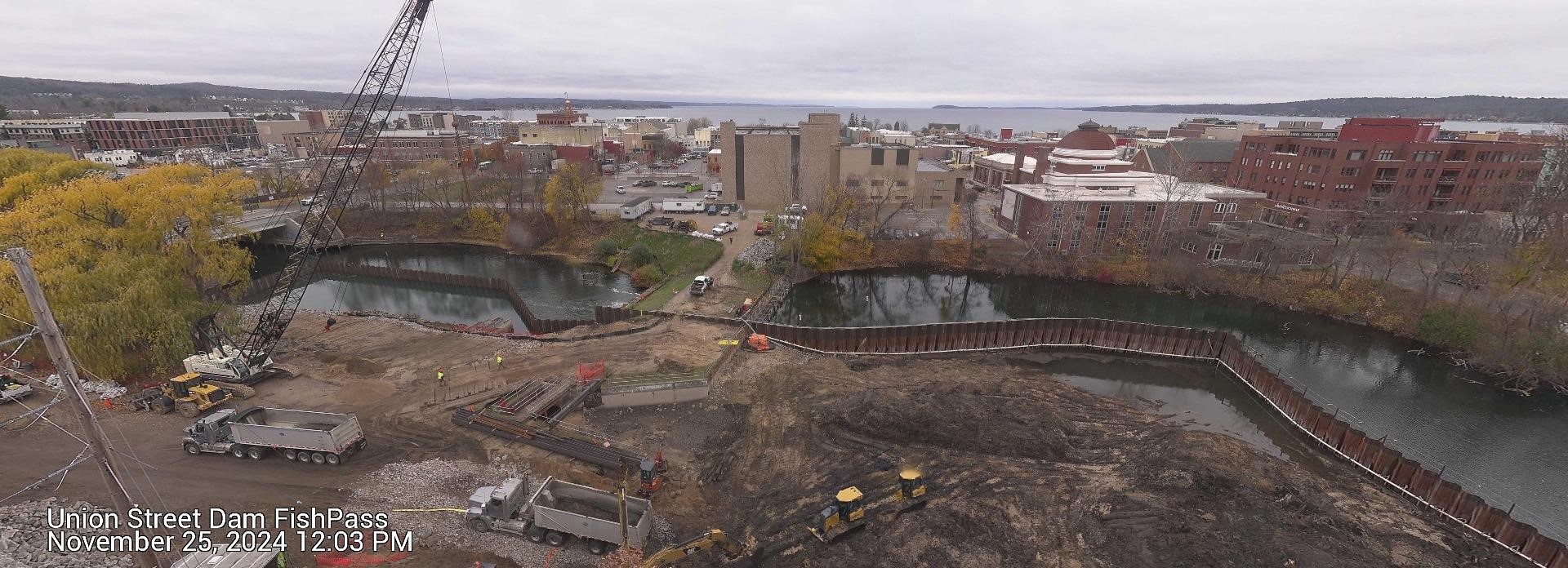 Aerial view of the Union Street Dam FishPass construction site taken on November 25, 2024, at 12:03 PM. The image shows continued construction progress with heavy equipment, including dump trucks and excavators, visible on the construction site. Sheet piling sections still divide parts of the river, and workers in high-visibility clothing are on-site. Surrounding the site are trees with late autumn foliage, urban buildings, and a view of the bay under a cloudy sky